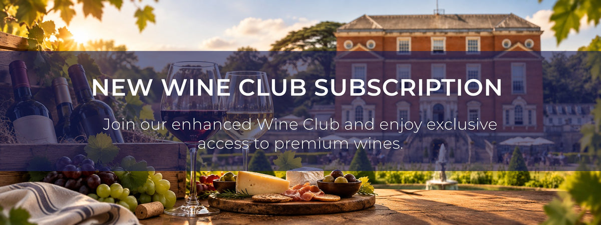 Promotional banner for a wine club subscription, featuring a blurred background of a grand estate building at sunset, with a foreground table styled with wine bottles, grapes, cheese, crackers, and a glass of red wine. White text across the center reads 'NEW WINE CLUB SUBSCRIPTION' and 'Join our enhanced Wine Club and enjoy exclusive access to premium wines.